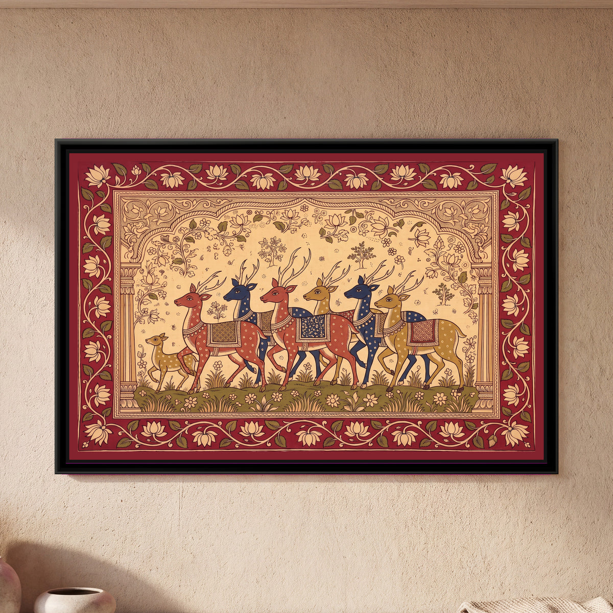 Kalamkari Painting – Procession of Spotted Deer - framed canvas print of six spotted deer with tall antlers in red, indigo and mustard walking left in a row, each wearing a patterned saddle cloth, a small spotted fawn leading at the front, a cream ground with lotus vines and small trees under a carved arch on two pillars, a green strip of grass and flowers below, an outer deep red border of cream lotuses on a trailing vine, hung on a wall