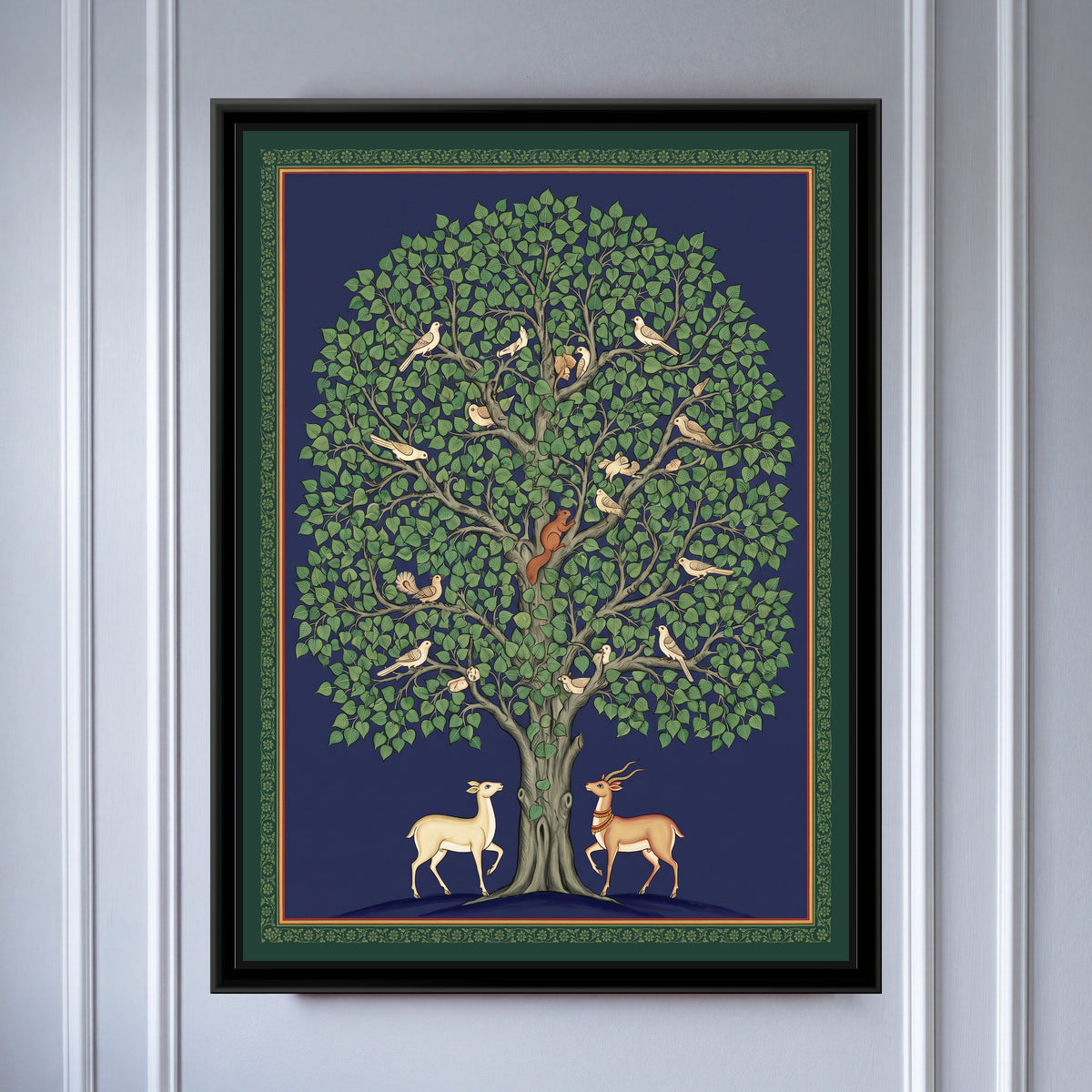 Pichwai Painting – Peepal Tree with Birds - framed canvas print of a large peepal tree with a dense dome of heart-shaped green leaves on a midnight blue ground, about twenty cream and tan birds perched along its grey branches, a red squirrel climbing the trunk, a pale doe and a tan blackbuck with spiral horns facing each other at the foot of the tree, a slim dark green border of tiny flowers with a thin red line, hung on a wall