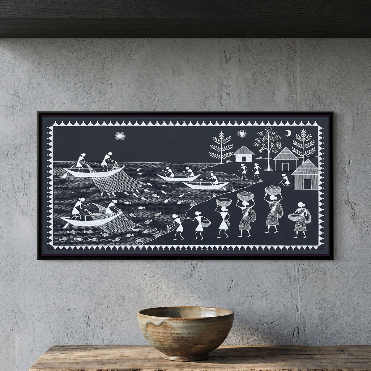Warli Painting – Fishermen on the River - framed canvas print of a Warli river scene in white line on charcoal: fishermen in long boats hauling nets full of fish, rowers on the water, a riverbank where women carry baskets on their heads, thatched huts and leafy trees beyond, sun and crescent moon, inside a border of white triangles, hung on a wall