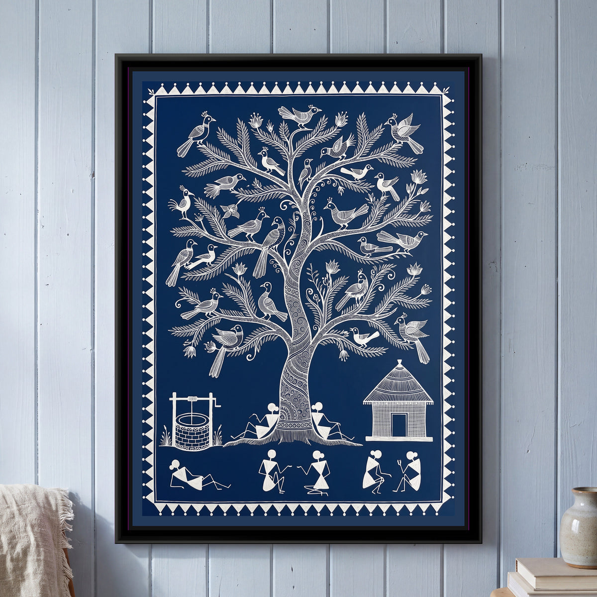Warli Painting – Tree of Life with Birds – Deep Indigo - framed canvas print of a tall Warli tree of life with a spiral-patterned trunk and feathery branches holding around thirty crested birds and small lotus buds, two villagers leaning against the trunk, a brick well with a winch on the left, a thatched hut on the right and a row of stick-figure villagers chatting and resting below, white line on a deep indigo ground inside a border of white triangles and dots, hung on a wall