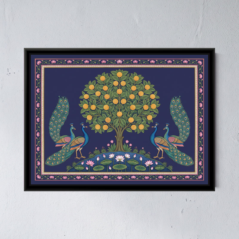 Pichwai Painting – Peacocks Flanking a Kadamba Tree - framed canvas print of a kadamba tree with a round crown of green leaves and round golden-orange blossoms standing on a small mound above a lotus pond, two pairs of blue and teal peacocks facing the tree from either side with their eyed tails raised and trailing, pink and white lotuses on green pads in the pond, a navy ground, a border of pink lotuses on a vine with a slim cream and gold inner band, hung on a wall