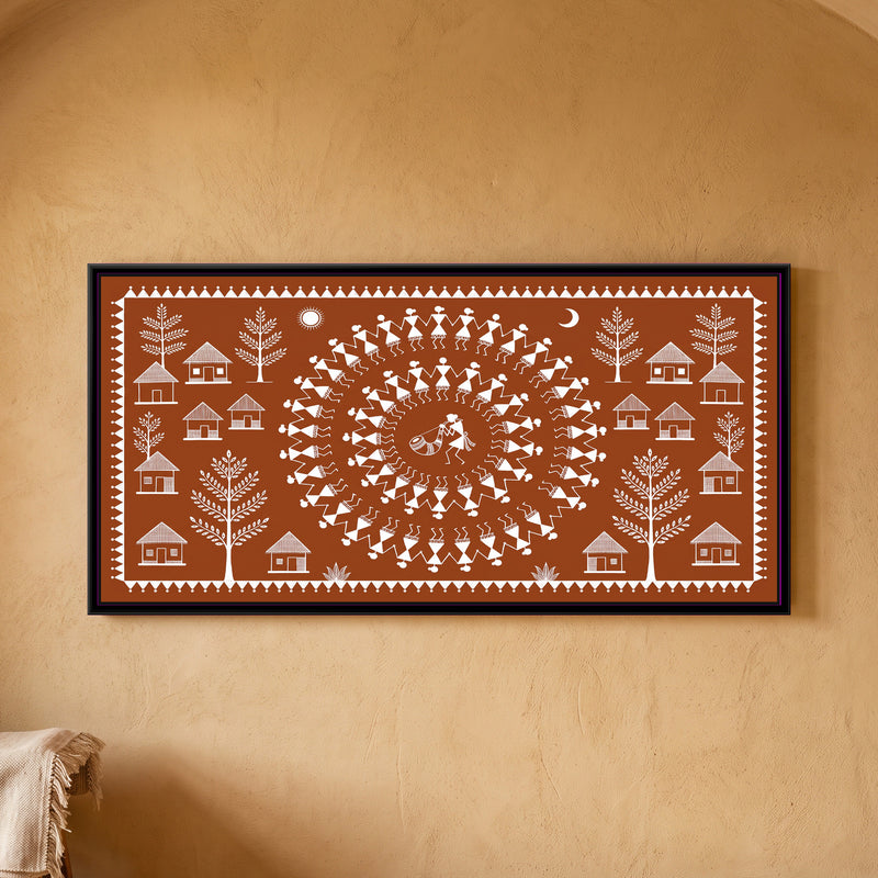 Warli Painting – Tarpa Dance Circle - framed canvas print of two concentric rings of white Warli stick-figure dancers holding hands around a tarpa horn player at the centre, thatched huts and leafy trees on both sides, a sun and a crescent moon above, white line on a terracotta ground inside a border of white triangles, hung on a wall