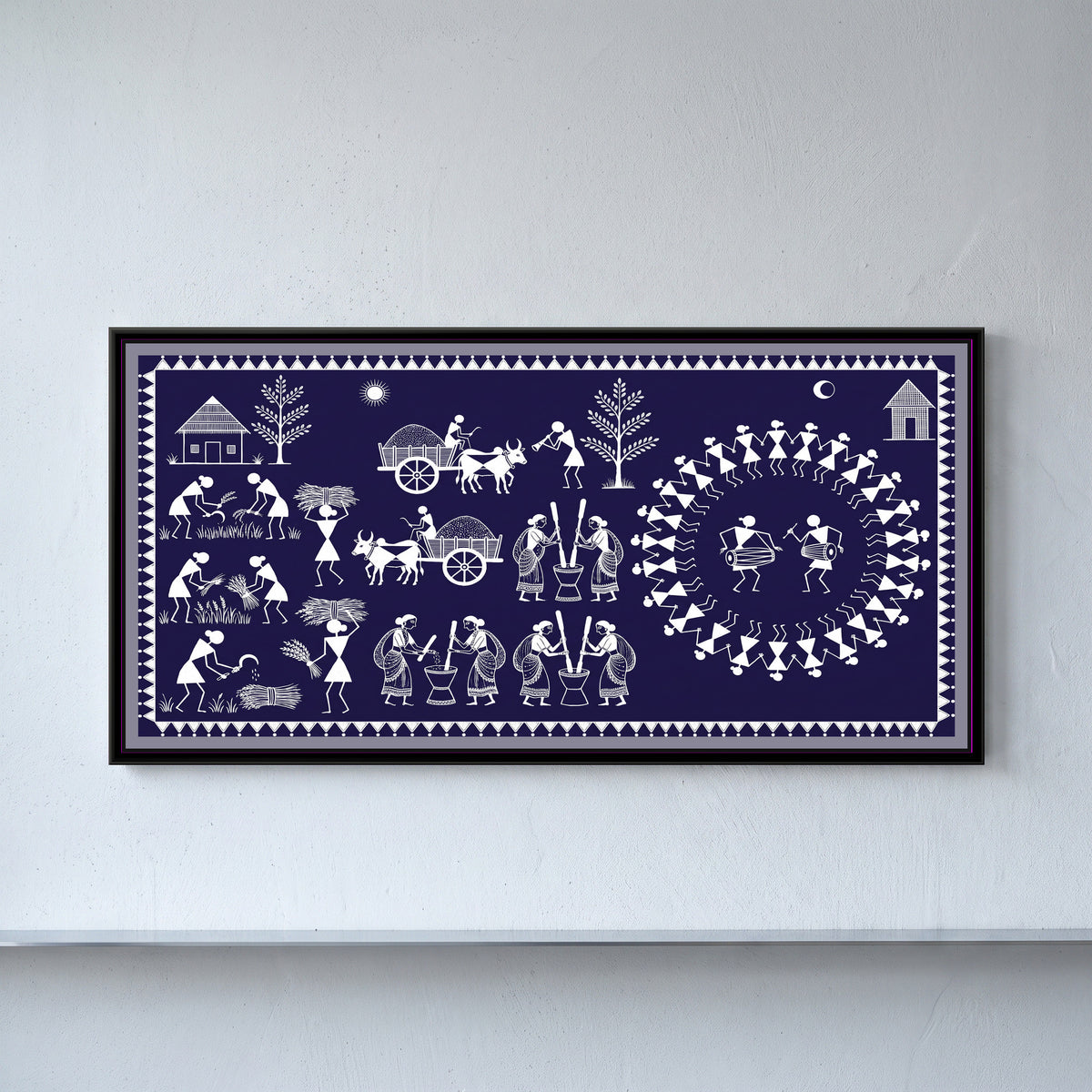 Warli Painting – Harvest Festival - framed canvas print of a Warli harvest scene in white line on deep indigo: villagers cutting and bundling grain with sickles, women pounding rice in mortars, two bullock carts loaded with grain, a horn player, a ring of dancers around two drummers, a hut, trees, sun and crescent moon, inside a border of white triangles, hung on a wall