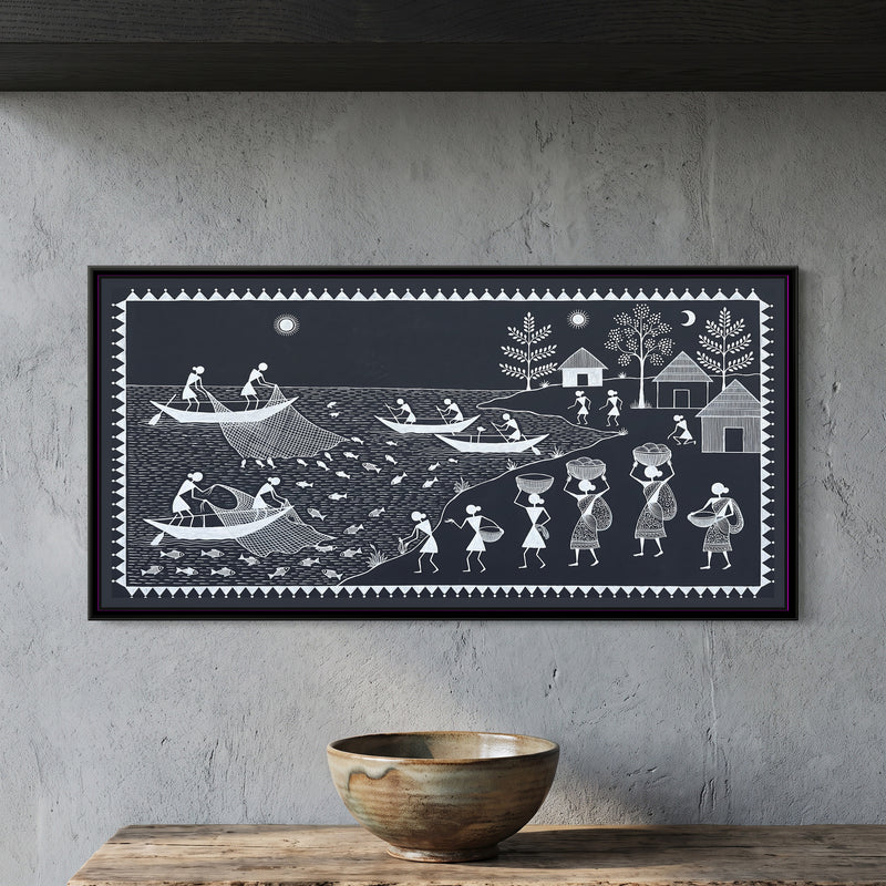 Warli Painting – Fishermen on the River - framed canvas print of a Warli river scene in white line on charcoal: fishermen in long boats hauling nets full of fish, rowers on the water, a riverbank where women carry baskets on their heads, thatched huts and leafy trees beyond, sun and crescent moon, inside a border of white triangles, hung on a wall