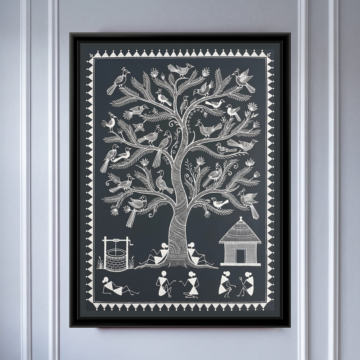 Warli Painting – Tree of Life with Birds – Deep Charcoal - framed canvas print of a tall Warli tree of life with a spiral-patterned trunk and feathery branches holding around thirty crested birds and small lotus buds, two villagers leaning against the trunk, a brick well with a winch on the left, a thatched hut on the right and a row of stick-figure villagers chatting and resting below, white line on a deep charcoal grey ground inside a border of white triangles and dots, hung on a wall